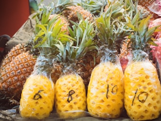 A collection of pineapples with some still in their natural form and others peeled and wrapped in plastic. The peeled pineapples have numbers written on them with a marker, indicating potential pricing or weight.