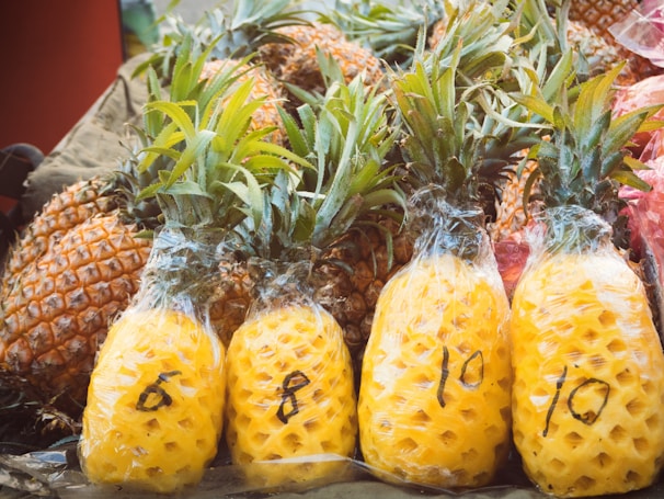 A collection of pineapples with some still in their natural form and others peeled and wrapped in plastic. The peeled pineapples have numbers written on them with a marker, indicating potential pricing or weight.