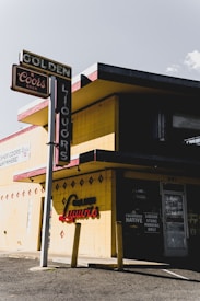 A vintage liquor store with a retro sign advertising Coors beer. The building facade is primarily yellow with red diamond-shaped accents and features bold signage reading 'Liquors'. There is a shadow cast by the building, creating a stark contrast between sunlight and shade.