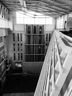 A modern architectural interior featuring large vertical pipes, industrial design elements, and expansive windows allowing natural light. The space features a high ceiling with visible steel beams and a large diagonal structure in the foreground.
