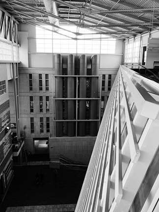 A modern architectural interior featuring large vertical pipes, industrial design elements, and expansive windows allowing natural light. The space features a high ceiling with visible steel beams and a large diagonal structure in the foreground.