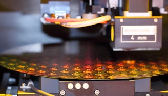 A close-up view of a semiconductor wafer being processed in a machine. The surface of the wafer displays intricate patterns of microchips with a reflective, multicolored appearance. The machine features various mechanical components, including cables and a label reading 'Writemode 4 mm'.