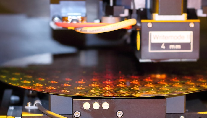 A close-up view of a semiconductor wafer being processed in a machine. The surface of the wafer displays intricate patterns of microchips with a reflective, multicolored appearance. The machine features various mechanical components, including cables and a label reading 'Writemode 4 mm'.