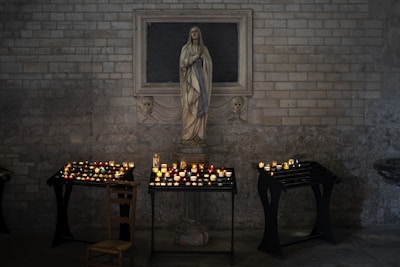A stone statue of a religious figure stands against a brick wall, surrounded by multiple small, lit candles on black stands. A wooden chair is placed nearby, and the setting has a dim, serene atmosphere.