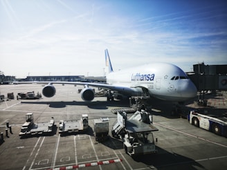 A large commercial airplane is stationed at an airport gate, with one side prominently displaying the Lufthansa logo. The aircraft is connected to a jet bridge, and various ground service vehicles are positioned around it, including cargo loaders and tugs. The sky is clear with a few scattered clouds and the airport area looks expansive.