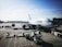 A large commercial airplane is stationed at an airport gate, with one side prominently displaying the Lufthansa logo. The aircraft is connected to a jet bridge, and various ground service vehicles are positioned around it, including cargo loaders and tugs. The sky is clear with a few scattered clouds and the airport area looks expansive.