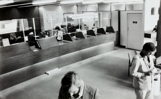 A black and white image of a bank's interior, showing a row of teller stations with glass dividers. Two employees are seated behind the counter and one is using a phone. Two men in suits are visible in the foreground, presumably customers, interacting with documents.