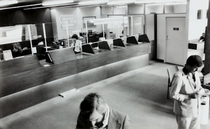 A black and white image of a bank's interior, showing a row of teller stations with glass dividers. Two employees are seated behind the counter and one is using a phone. Two men in suits are visible in the foreground, presumably customers, interacting with documents.