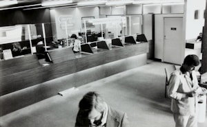 A black and white image of a bank's interior, showing a row of teller stations with glass dividers. Two employees are seated behind the counter and one is using a phone. Two men in suits are visible in the foreground, presumably customers, interacting with documents.