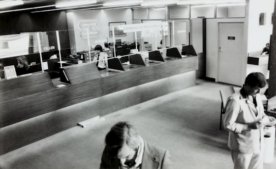 A black and white image of a bank's interior, showing a row of teller stations with glass dividers. Two employees are seated behind the counter and one is using a phone. Two men in suits are visible in the foreground, presumably customers, interacting with documents.