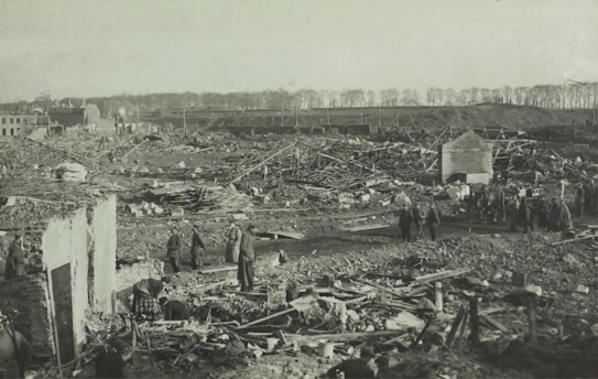 A vast landscape of destruction shows the aftermath of a significant event. The ground is littered with debris and remnants of structures. People are scattered across the scene, appearing to survey the damage or search through the rubble. In the background, there are partial ruins of buildings and a line of trees, suggesting a rural or semi-rural setting. Horses and carts are also visible, indicating a historical setting.