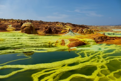 A striking landscape with vibrant yellow and green mineral deposits on the ground, surrounding pools of clear water. Rocky formations are scattered throughout the scene under a clear blue sky, creating a surreal and otherworldly appearance.
