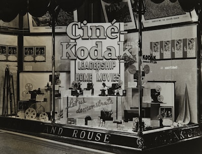 A vintage store display featuring Cine-Kodak cameras and equipment. The window display includes camera models, film reels, and promotional materials. Large, bold signage emphasizes leadership in home movies. Posters and framed images adorn the background.