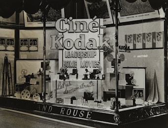 A vintage store display featuring Cine-Kodak cameras and equipment. The window display includes camera models, film reels, and promotional materials. Large, bold signage emphasizes leadership in home movies. Posters and framed images adorn the background.