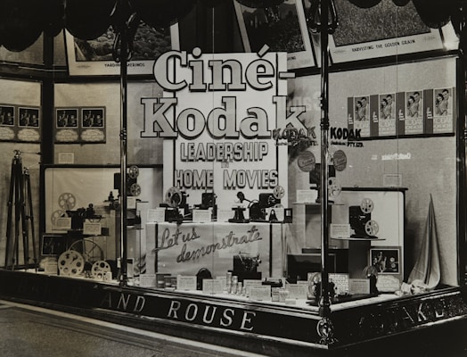 A vintage store display featuring Cine-Kodak cameras and equipment. The window display includes camera models, film reels, and promotional materials. Large, bold signage emphasizes leadership in home movies. Posters and framed images adorn the background.