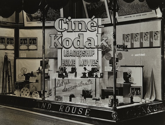 A vintage store display featuring Cine-Kodak cameras and equipment. The window display includes camera models, film reels, and promotional materials. Large, bold signage emphasizes leadership in home movies. Posters and framed images adorn the background.