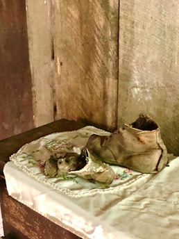 A pair of worn, vintage leather shoes rest on an embroidered cloth placed on a wooden surface. The background features a textured, wooden wall, creating a rustic and nostalgic atmosphere.