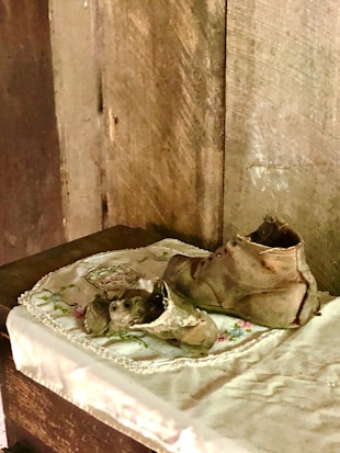 A pair of worn, vintage leather shoes rest on an embroidered cloth placed on a wooden surface. The background features a textured, wooden wall, creating a rustic and nostalgic atmosphere.