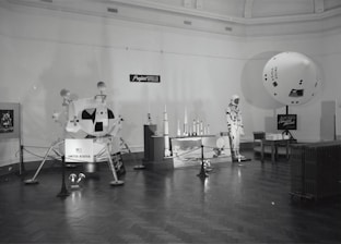 A museum exhibit features various models and displays related to Project Apollo. Central to the exhibit is a model of a Lunar Module labeled 'United States.' There are scale models of rockets on a platform, a mannequin dressed in an astronaut suit, and an informational board about Apollo Command and Models. The exhibit is arranged in a spacious room with wood flooring and a high ceiling.