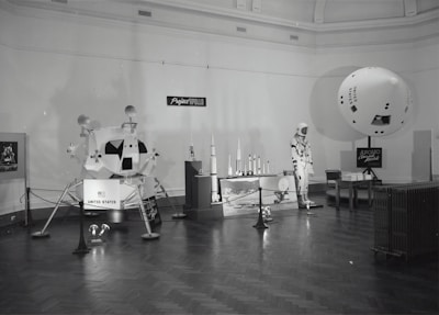 A museum exhibit features various models and displays related to Project Apollo. Central to the exhibit is a model of a Lunar Module labeled 'United States.' There are scale models of rockets on a platform, a mannequin dressed in an astronaut suit, and an informational board about Apollo Command and Models. The exhibit is arranged in a spacious room with wood flooring and a high ceiling.