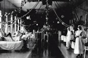 A large hospital ward filled with beds, where several patients are either sitting or lying down. Two nurses in white uniforms are standing to the right, attending to patients. The room is decorated with garlands and paper lanterns, suggesting a festive or ceremonial atmosphere.