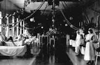 A large hospital ward filled with beds, where several patients are either sitting or lying down. Two nurses in white uniforms are standing to the right, attending to patients. The room is decorated with garlands and paper lanterns, suggesting a festive or ceremonial atmosphere.