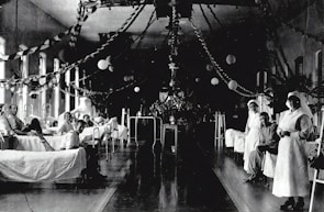 A large hospital ward filled with beds, where several patients are either sitting or lying down. Two nurses in white uniforms are standing to the right, attending to patients. The room is decorated with garlands and paper lanterns, suggesting a festive or ceremonial atmosphere.
