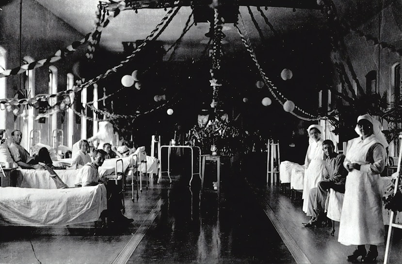 A large hospital ward filled with beds, where several patients are either sitting or lying down. Two nurses in white uniforms are standing to the right, attending to patients. The room is decorated with garlands and paper lanterns, suggesting a festive or ceremonial atmosphere.