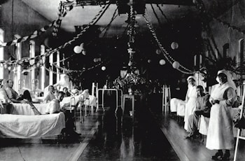A large hospital ward filled with beds, where several patients are either sitting or lying down. Two nurses in white uniforms are standing to the right, attending to patients. The room is decorated with garlands and paper lanterns, suggesting a festive or ceremonial atmosphere.