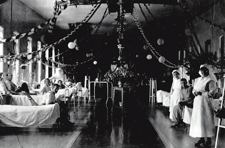 A large hospital ward filled with beds, where several patients are either sitting or lying down. Two nurses in white uniforms are standing to the right, attending to patients. The room is decorated with garlands and paper lanterns, suggesting a festive or ceremonial atmosphere.