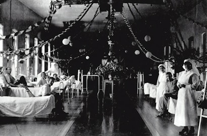 A large hospital ward filled with beds, where several patients are either sitting or lying down. Two nurses in white uniforms are standing to the right, attending to patients. The room is decorated with garlands and paper lanterns, suggesting a festive or ceremonial atmosphere.