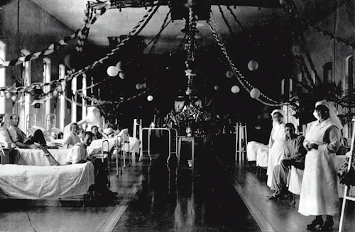 A large hospital ward filled with beds, where several patients are either sitting or lying down. Two nurses in white uniforms are standing to the right, attending to patients. The room is decorated with garlands and paper lanterns, suggesting a festive or ceremonial atmosphere.