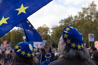 A crowd of people is gathered, many wearing blue berets adorned with yellow stars. Large blue flags with similar yellow stars are prominently displayed, suggesting a theme related to the European Union. The setting appears to be outdoors with trees visible in the background.