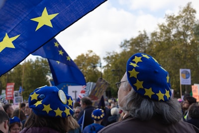 A crowd of people is gathered, many wearing blue berets adorned with yellow stars. Large blue flags with similar yellow stars are prominently displayed, suggesting a theme related to the European Union. The setting appears to be outdoors with trees visible in the background.