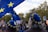 A crowd of people is gathered, many wearing blue berets adorned with yellow stars. Large blue flags with similar yellow stars are prominently displayed, suggesting a theme related to the European Union. The setting appears to be outdoors with trees visible in the background.