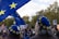 A crowd of people is gathered, many wearing blue berets adorned with yellow stars. Large blue flags with similar yellow stars are prominently displayed, suggesting a theme related to the European Union. The setting appears to be outdoors with trees visible in the background.