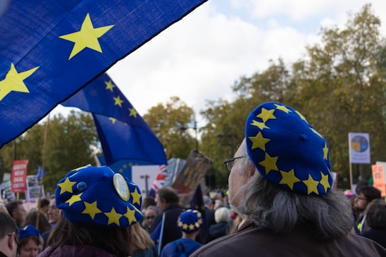 A crowd of people is gathered, many wearing blue berets adorned with yellow stars. Large blue flags with similar yellow stars are prominently displayed, suggesting a theme related to the European Union. The setting appears to be outdoors with trees visible in the background.