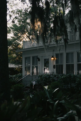 Twilight view of a historic home with warm lights glowing inside and Spanish moss hanging from nearby trees.