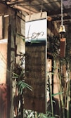A wooden signboard hanging in a rustic setting, with handwritten text listing items such as handmade crafts, clothes, accessories, wind chimes, books, and pots & plants. The background includes bamboo and other wooden structures, giving a cozy and natural ambiance.