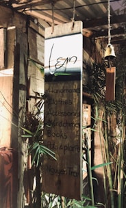 A wooden signboard hanging in a rustic setting, with handwritten text listing items such as handmade crafts, clothes, accessories, wind chimes, books, and pots & plants. The background includes bamboo and other wooden structures, giving a cozy and natural ambiance.