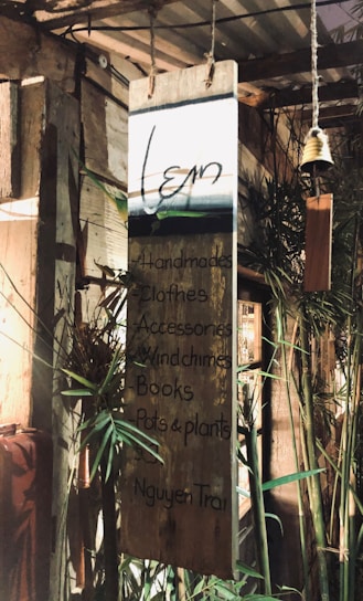 A wooden signboard hanging in a rustic setting, with handwritten text listing items such as handmade crafts, clothes, accessories, wind chimes, books, and pots & plants. The background includes bamboo and other wooden structures, giving a cozy and natural ambiance.
