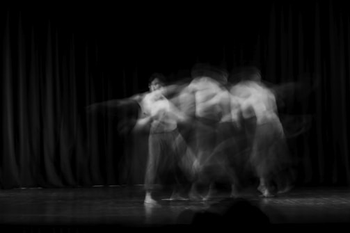 A black and white long-exposure photograph capturing the blurred motion of a dancer on stage. The figure appears multiple times across the frame due to motion blur, suggesting dynamic movement.