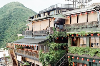 A multi-level building adorned with vibrant greenery and traditional red lanterns, set against a lush mountainous backdrop. The structure features wooden accents and open balconies, creating a harmonious blend with nature.