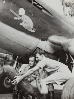 Two men dressed in military-style clothing are handling a large cylindrical object under the wing of a vintage aircraft. The aircraft features distinctive nose art depicting a child-like figure in pajamas. The atmosphere suggests a wartime setting, possibly during a preparation or maintenance period.