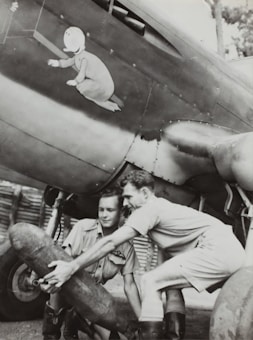 Two men dressed in military-style clothing are handling a large cylindrical object under the wing of a vintage aircraft. The aircraft features distinctive nose art depicting a child-like figure in pajamas. The atmosphere suggests a wartime setting, possibly during a preparation or maintenance period.