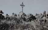 A group of soldiers in military uniforms and wide-brimmed hats are gathered around a wooden cross in an outdoor setting. The grass and vegetation suggest a rural or battlefield location. The scene appears somber, possibly a memorial or remembrance ceremony.