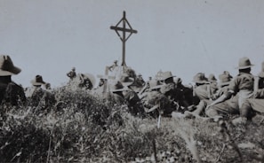 A group of soldiers in military uniforms and wide-brimmed hats are gathered around a wooden cross in an outdoor setting. The grass and vegetation suggest a rural or battlefield location. The scene appears somber, possibly a memorial or remembrance ceremony.