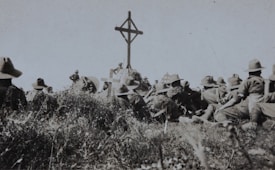 A group of soldiers in military uniforms and wide-brimmed hats are gathered around a wooden cross in an outdoor setting. The grass and vegetation suggest a rural or battlefield location. The scene appears somber, possibly a memorial or remembrance ceremony.