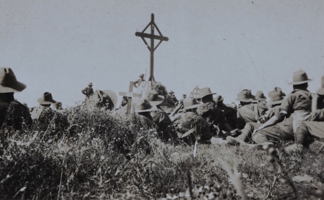 A group of soldiers in military uniforms and wide-brimmed hats are gathered around a wooden cross in an outdoor setting. The grass and vegetation suggest a rural or battlefield location. The scene appears somber, possibly a memorial or remembrance ceremony.