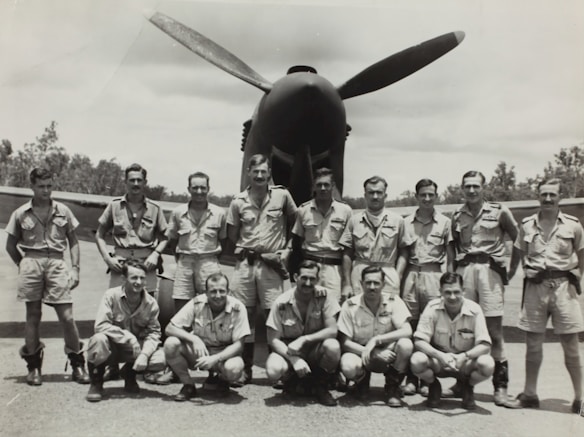 A group of men in military uniforms are posing in front of an aircraft. They appear to be part of a World War II era flight crew, standing and crouching in front of the plane with a propeller prominent in the background. The men are dressed in short-sleeved shirts and shorts, typical of tropical or warm climate uniforms. The photo is black and white, indicating an older photograph. Trees and a clear sky are visible in the background.